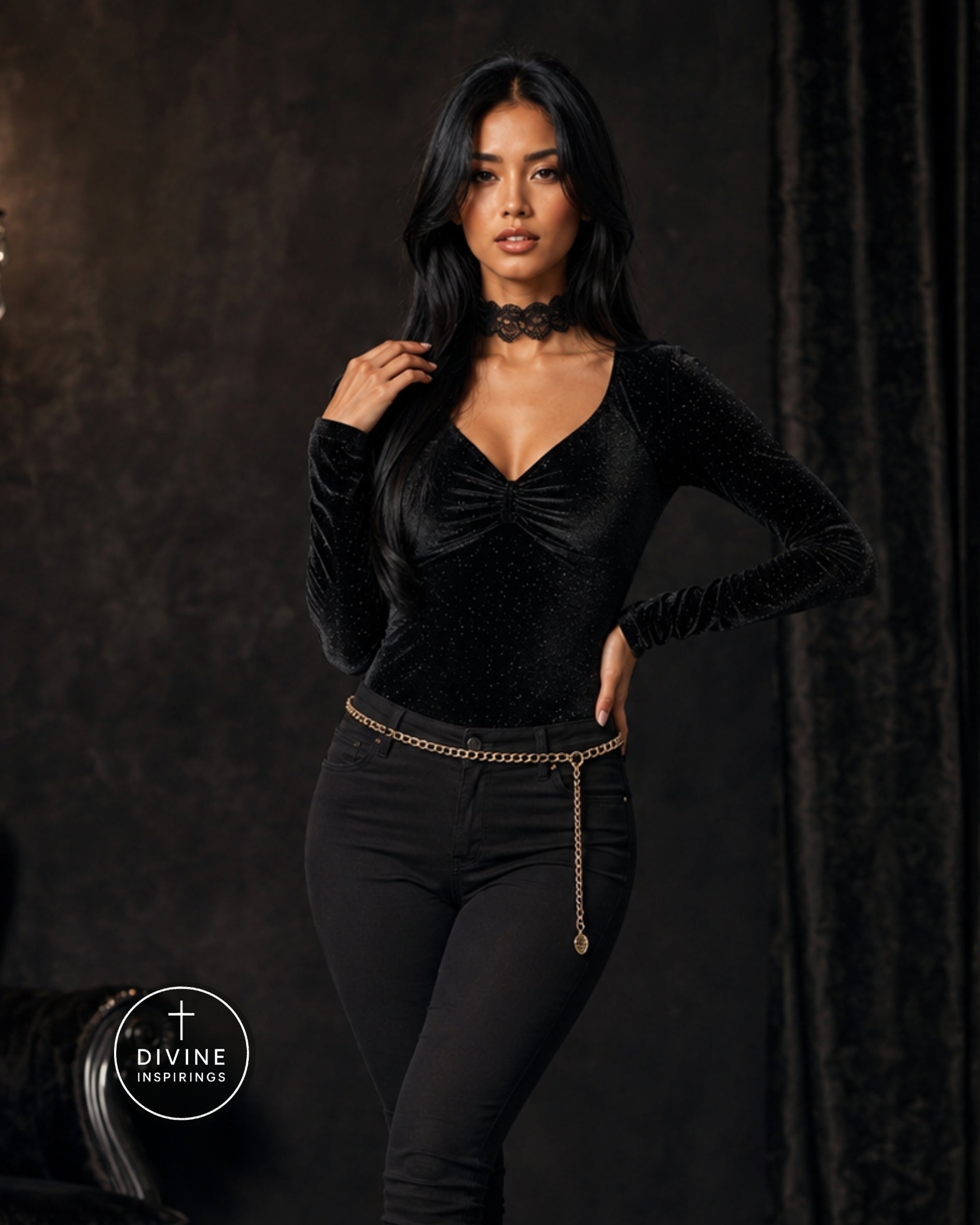 A woman modeling a black long-sleeve velvet top with a ruched sweetheart neckline and a black choker, paired with black pants and a gold chain belt, against a dark textured studio backdrop.