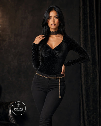 A woman modeling a black long-sleeve velvet top with a ruched sweetheart neckline and a black choker, paired with black pants and a gold chain belt, against a dark textured studio backdrop.