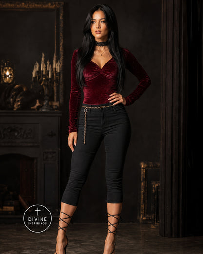 A woman models a gothic-style burgundy velvet long-sleeve top with a ruched sweetheart neckline and black skinny capri pants featuring a gold chain detail at the waist, set against a dark, ornate interior background.