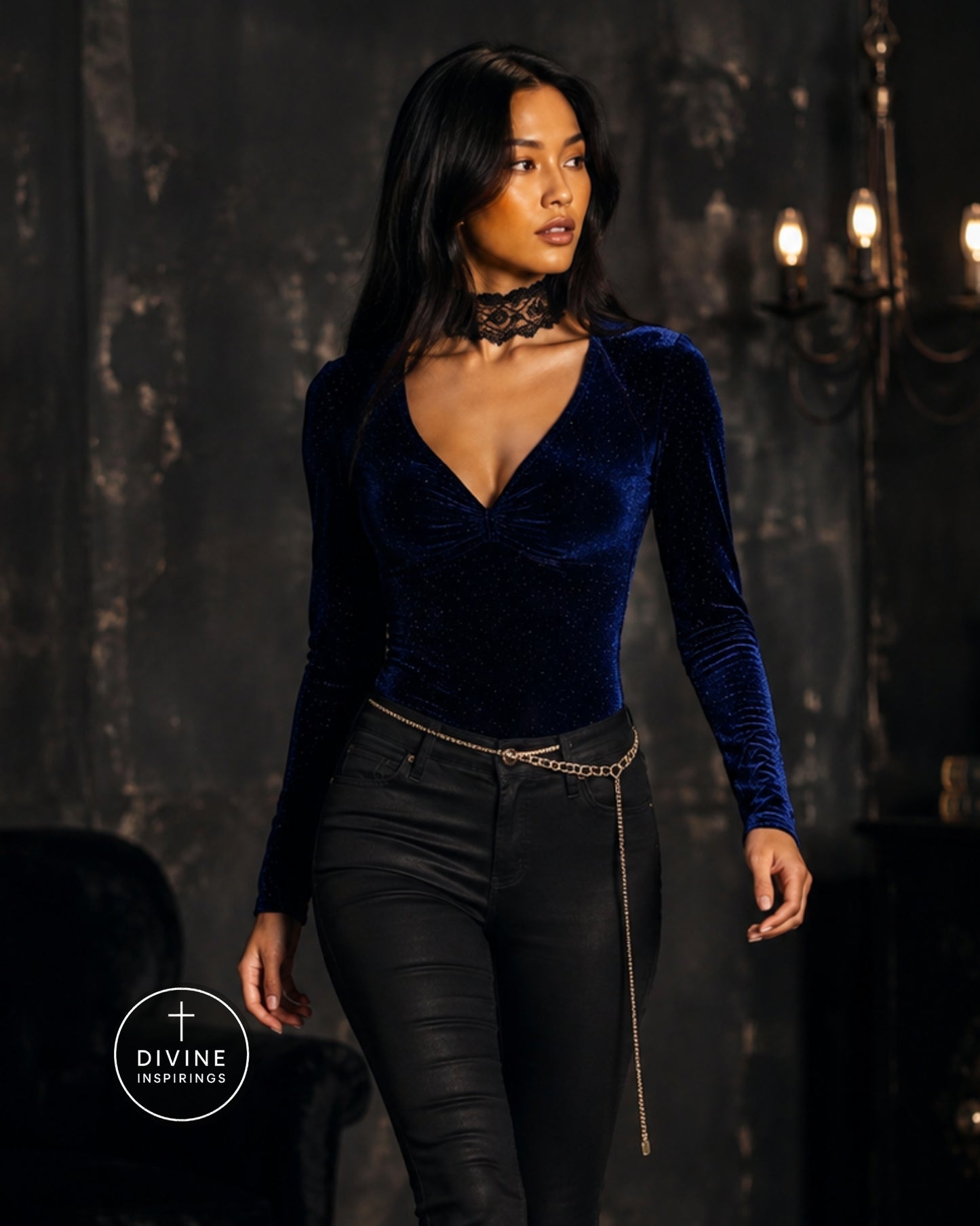 A woman modeling a dark blue velvet long-sleeve wrap-style top with black skinny pants and a gold chain belt, standing in a dimly lit gothic interior. A Divine Inspirings logo is visible in the bottom-left corner.