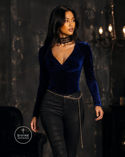 A woman modeling a dark blue velvet long-sleeve wrap-style top with black skinny pants and a gold chain belt, standing in a dimly lit gothic interior. A Divine Inspirings logo is visible in the bottom-left corner.