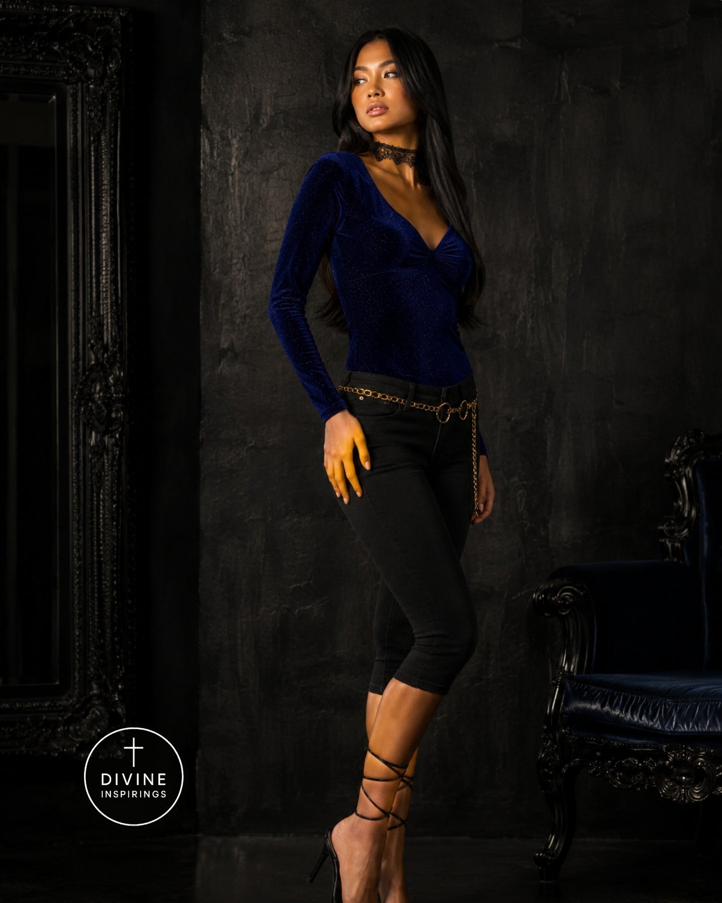 A woman models a dark blue velvet long-sleeve top with a deep V-neck, paired with black capri pants and gold accessories, set against a dark textured studio background with a mirror and an armchair.