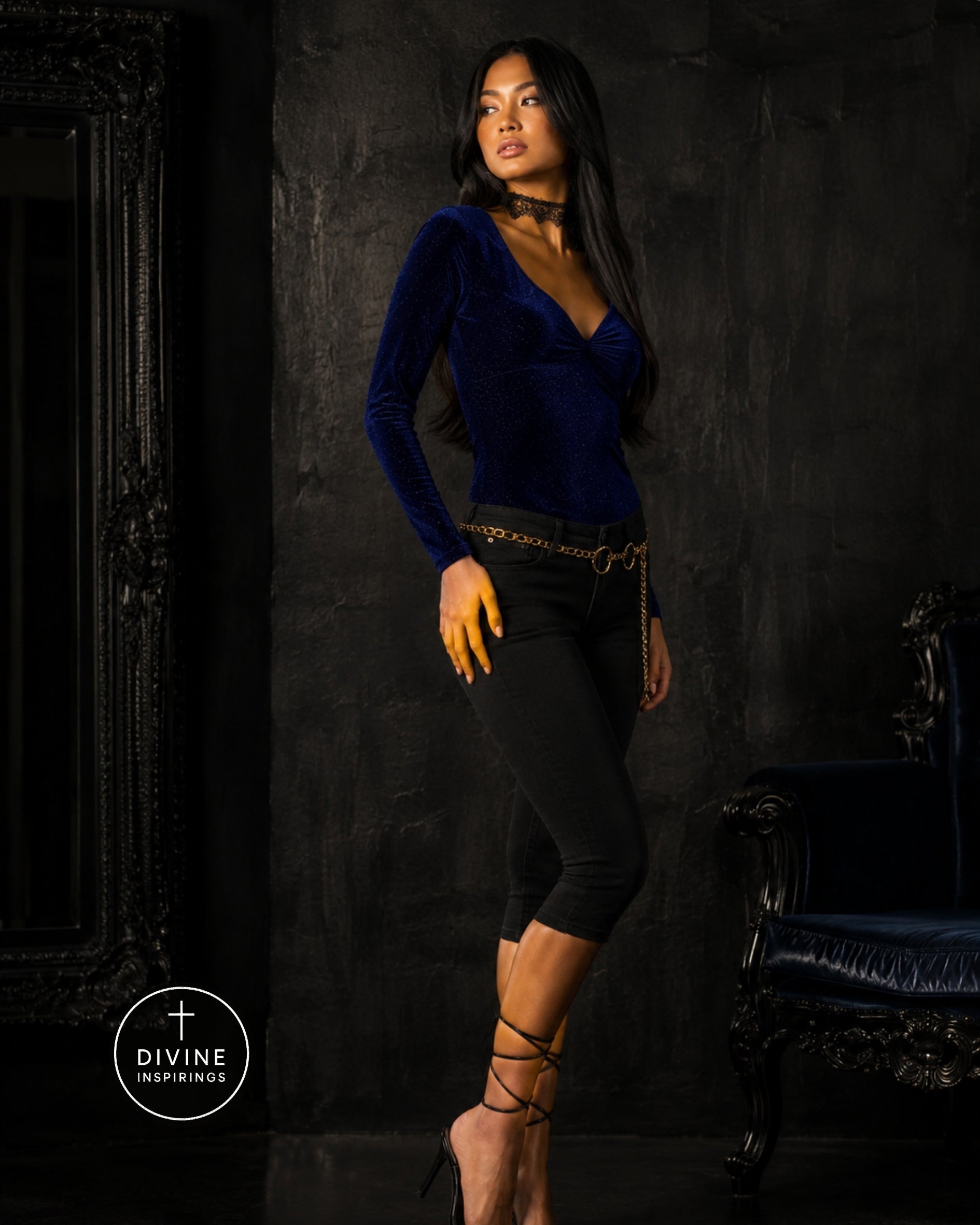 A woman models a dark blue velvet long-sleeve top with a deep V-neck, paired with black capri pants and gold accessories, set against a dark textured studio background with a mirror and an armchair.