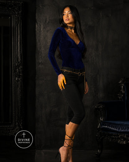 A woman models a dark blue velvet long-sleeve top with a deep V-neck, paired with black capri pants and gold accessories, set against a dark textured studio background with a mirror and an armchair.