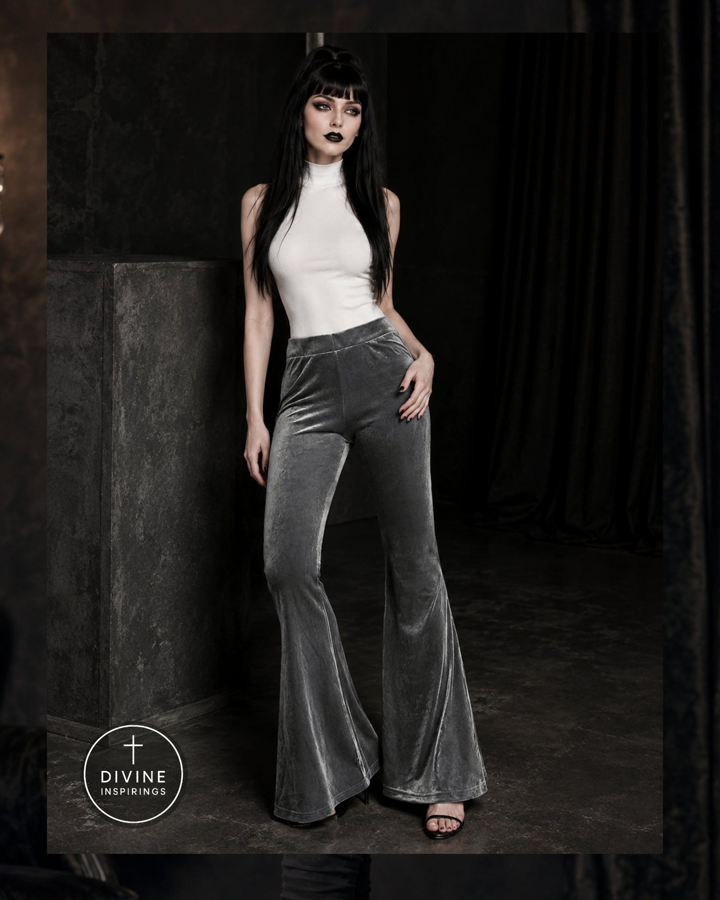 A woman modeling high-waisted, wide-leg velvet flare pants in a dark gray color, paired with a white sleeveless top, in a moody studio setting with a dark curtain background.