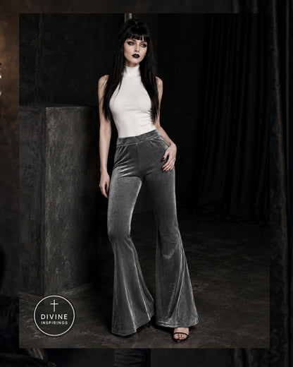 A woman modeling high-waisted, wide-leg velvet flare pants in a dark gray color, paired with a white sleeveless top, in a moody studio setting with a dark curtain background.