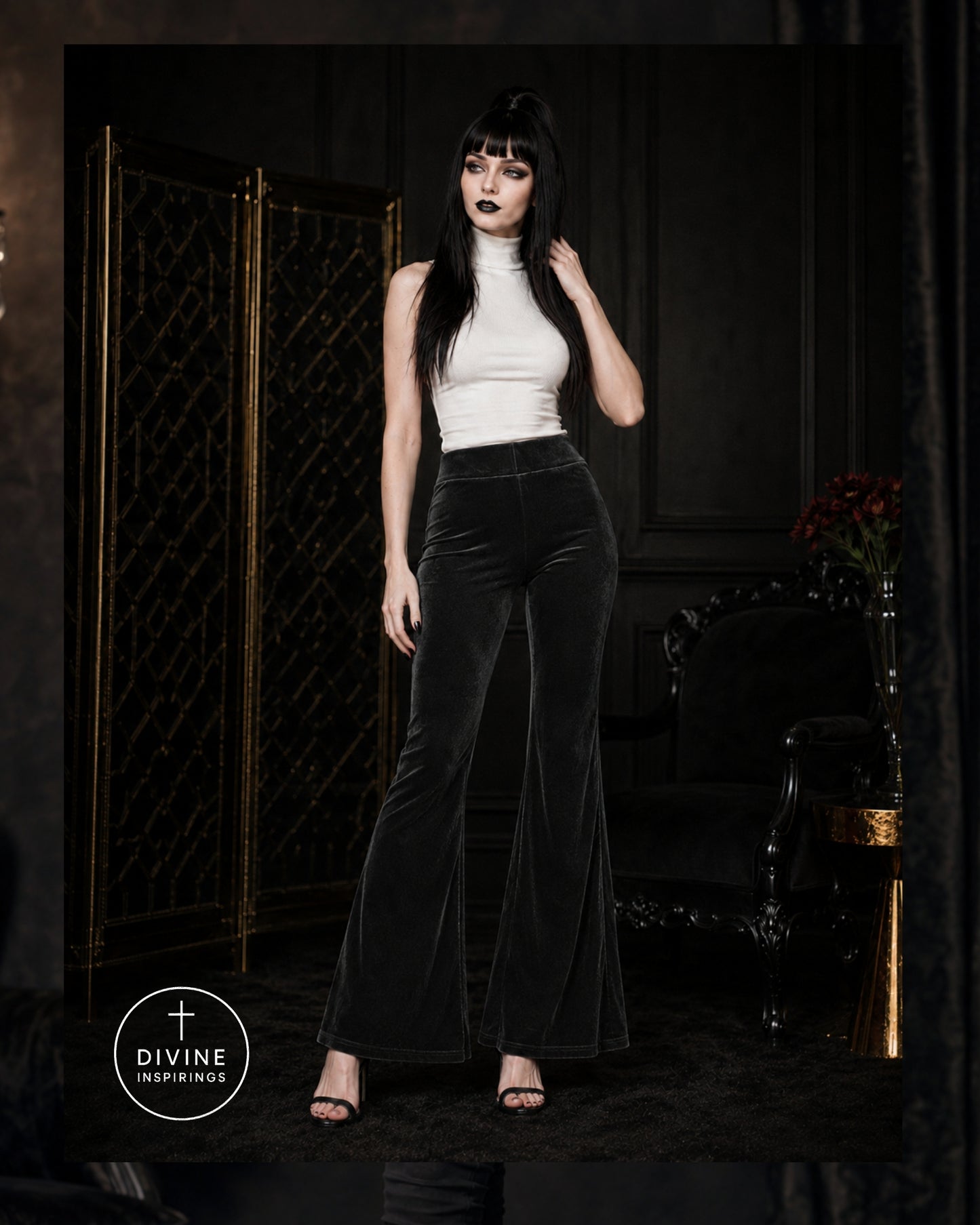 A woman modeling high-waisted black velvet flare pants with a white mock-neck top in a dark, gothic-themed room with a gold folding screen.