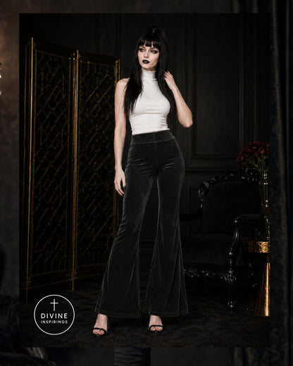 A woman modeling high-waisted black velvet flare pants with a white mock-neck top in a dark, gothic-themed room with a gold folding screen.