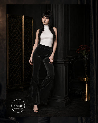 A model with long black hair and dark makeup stands in a dark interior wearing a white sleeveless mock-neck top and black velvet flare pants. A circular 'DIVINE INSPIRINGS' logo is visible in the lower-left corner.