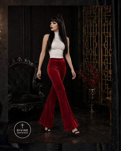 A woman models a two-piece outfit consisting of a white sleeveless mock-neck top and high-waisted, red velvet flare pants. She is standing in a dark, elegant room with a black velvet armchair and a gold decorative screen behind her.