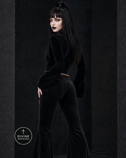 A model with long black hair and dark makeup poses in a black velvet jumpsuit with a gold chain detail at the waist, set against a dark textured studio background.