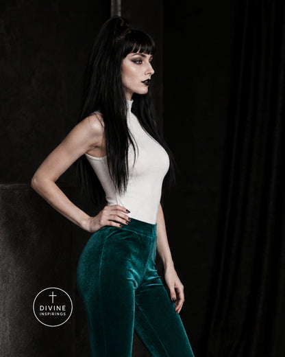 A woman posing in a white mock-neck top and high-waisted emerald green velvet pants against a dark studio backdrop. A circular 'DIVINE INSPIRINGS' logo is visible in the bottom-left corner.