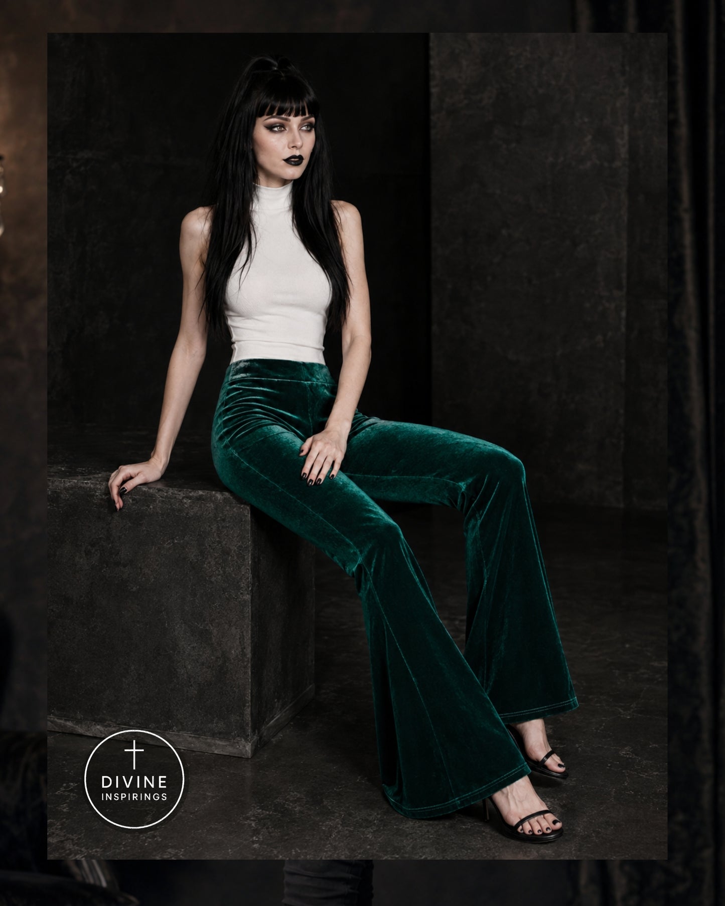 A model with long black hair and dark makeup sits on a dark stone block, wearing a white sleeveless mock-neck top and high-waisted, wide-leg emerald green velvet flare pants. The background is a dark, moody studio setting with textured stone walls.