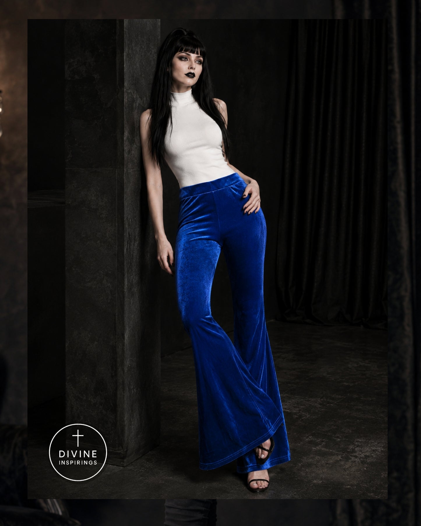 A model wearing a white mock-neck top and vibrant royal blue velvet flared pants, posing in a dark, moody studio setting with a visible 'DIVINE INSPIRINGS' logo in the bottom-left corner.
