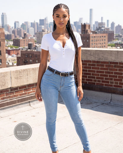 A woman stands on a rooftop wearing a white short-sleeve henley shirt and light blue skinny jeans with a black studded belt. The background shows a city skyline under a bright sky.
