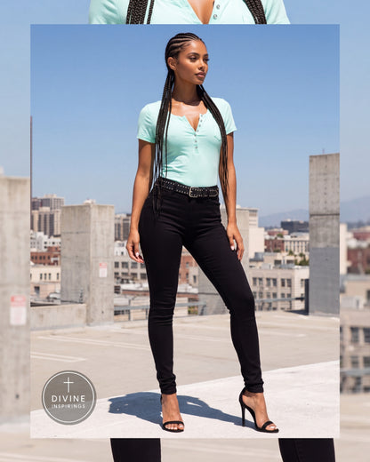A woman stands on a rooftop wearing a mint green short-sleeve henley top, black skinny jeans, a black belt, and black high-heeled sandals. The background shows a city skyline under a blue sky.