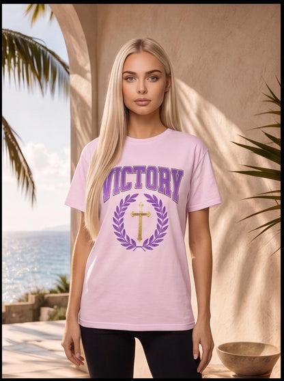 Woman wearing a pink t-shirt with 'VICTORY' and a cross design, standing outdoors with palm trees and ocean in the background.