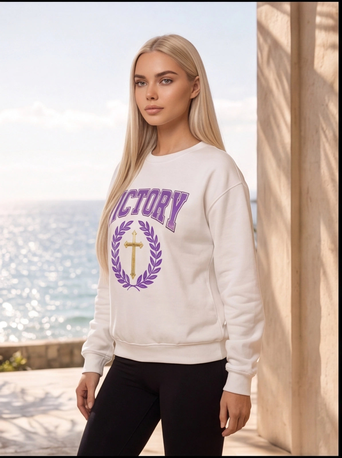 Woman wearing a white sweatshirt with 'VICTORY' and a cross design by the ocean.