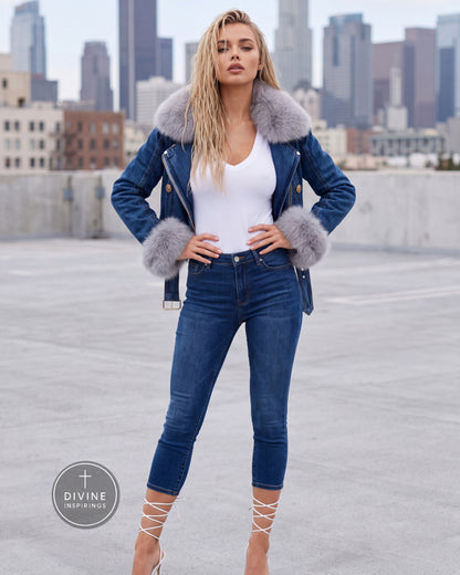 A woman stands on a rooftop wearing a blue denim jacket with a large gray faux-fur collar and cuffs, a white top, and blue skinny jeans. A city skyline is visible in the background, and a circular 'DIVINE INSPIRINGS' logo appears in the lower-left corner.