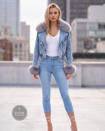 A woman stands on a rooftop wearing a light blue denim jacket with a large gray faux-fur collar and matching gray faux-fur trim at the cuffs, paired with light blue skinny jeans, a white top, and lace-up heels. A circular 'DIVINE INSPIRINGS' logo watermark is visible in the lower-left corner.
