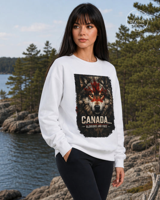 A woman wearing a white crewneck sweatshirt with a large graphic of a red-eyed wolf and the text 'CANADA GLORIOUS AND FREE' stands outdoors near a rocky shoreline and water. A circular 'DIVINE INSPIRINGS' logo is visible in the top right corner.