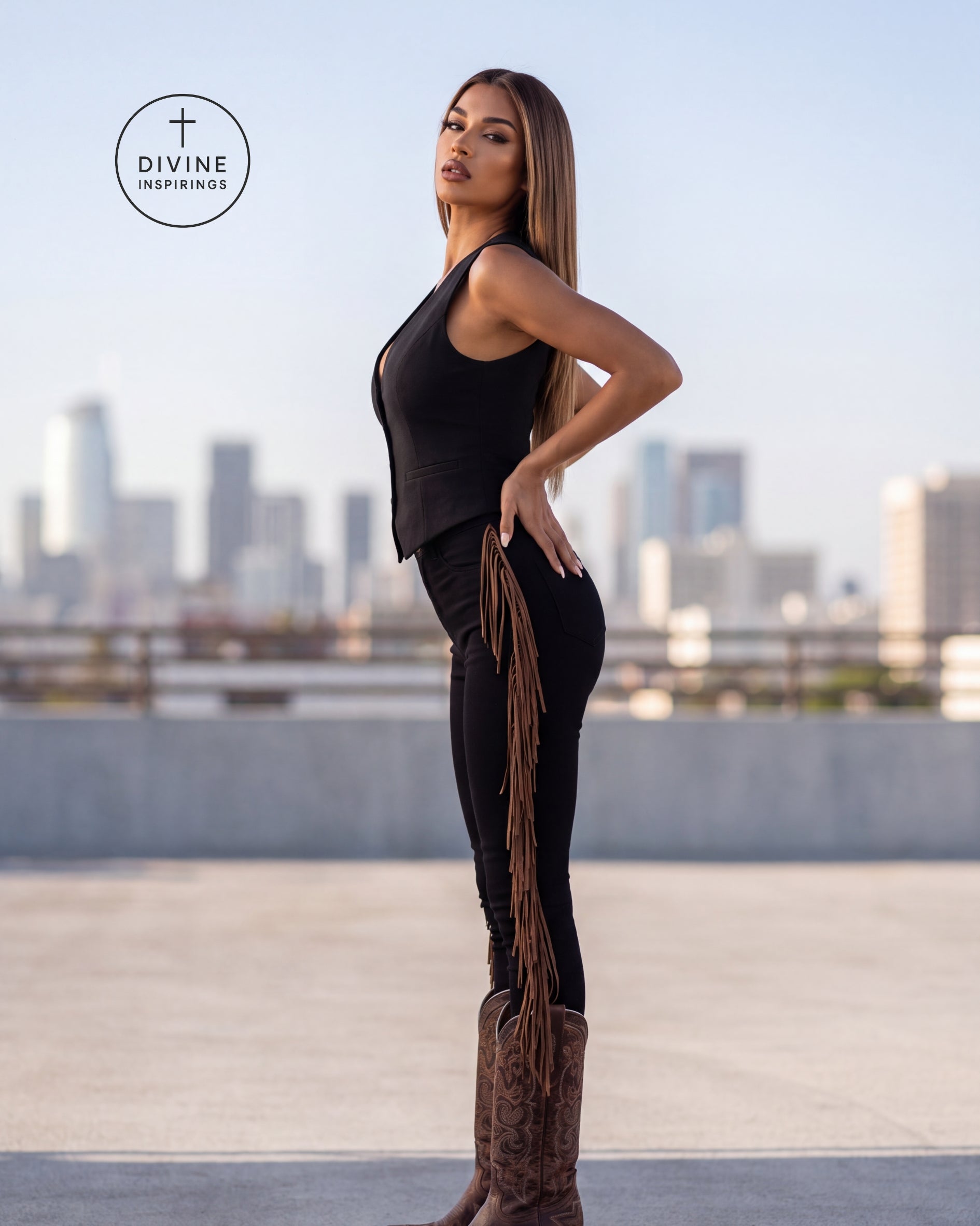 A woman stands on a rooftop wearing a black sleeveless top and black pants with a long fringe detail, paired with brown cowboy boots. The city skyline is blurred in the background, and a circular 'DIVINE INSPIRINGS' logo is visible in the upper left.