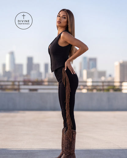 A woman stands on a rooftop wearing a black sleeveless top and black pants with a long fringe detail, paired with brown cowboy boots. The city skyline is blurred in the background, and a circular 'DIVINE INSPIRINGS' logo is visible in the upper left.