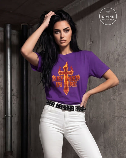 A woman wearing a purple Tee with a red and orange graphic that reads 'REFINED IN FIRE' under a flaming cross, paired with white pants and a gold chain belt, leaning against a concrete pillar.
