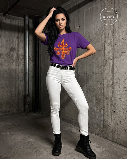 A woman wearing a purple tee with a large orange and yellow flame-cross graphic, "Refined in Fire" on the chest, paired with white fitted pants and a gold chain belt, standing against a dark concrete wall. A 'DIVINE INSPIRINGS' logo is visible in the top right corner.