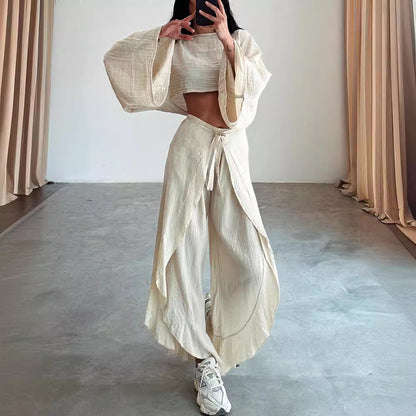 Person wearing a beige outfit with wide-legged pants and a top, standing in a minimalistic room with beige curtains.